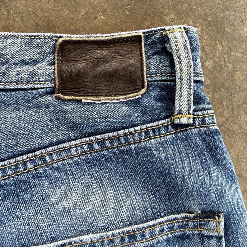 Pu&ograve; includere: Primo piano di jeans di denim blu con una toppa in pelle marrone. I jeans hanno cuciture gialle e un classico design a cinque tasche. Il denim ha un aspetto sbiadito e usurato.
