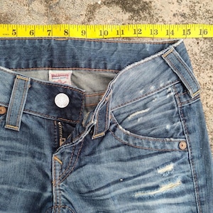 May include: Close-up of blue denim jeans with a button closure and zipper. The jeans have distressed details and a faded wash. A yellow measuring tape is placed across the top of the jeans, showing measurements from 12.7 to 40.6 cm.