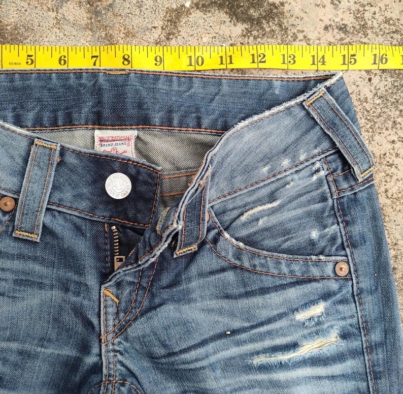 May include: Close-up of blue denim jeans with a button closure and zipper. The jeans have distressed details and a faded wash. A yellow measuring tape is placed across the top of the jeans, showing measurements from 12.7 to 40.6 cm.