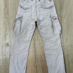 May include: Light beige cargo pants with multiple pockets and decorative stitching. The pants are hanging on a wooden hanger against a wooden background. The pants have a relaxed fit and a distressed look.