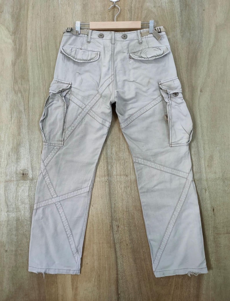May include: Light beige cargo pants with multiple pockets and decorative stitching. The pants are hanging on a wooden hanger against a wooden background. The pants have a relaxed fit and a distressed look.