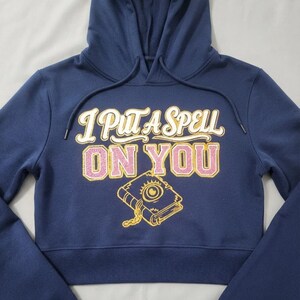 May include: A navy blue cropped hoodie with a drawstring hood. The front features the text "I PUT A SPELL ON YOU" in white and pink glittery lettering, accompanied by a graphic illustration of a closed book with an eye and a chain in gold.