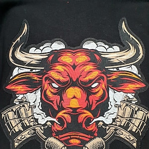 May include: Black crewneck sweatshirt featuring a detailed, embroidered bull design. The bull's head is rendered in shades of red, orange, and yellow, with white accents and stylized horns. Two tattoo machines are on either side of the bull's head.