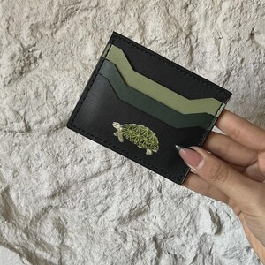 Puede incluir: Tarjetero de cuero negro con tres ranuras para tarjetas en tonos verdes. Un pequeño y detallado emblema de tortuga está en la parte delantera. El tarjetero se sostiene contra un fondo texturizado blanquecino.