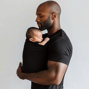 Peut inclure: Un porte-b&eacute;b&eacute; noir enveloppe un nourrisson endormi. Le porte-b&eacute;b&eacute; est tenu par une personne portant un t-shirt noir. L'arri&egrave;re-plan est un mur blanc uni.
