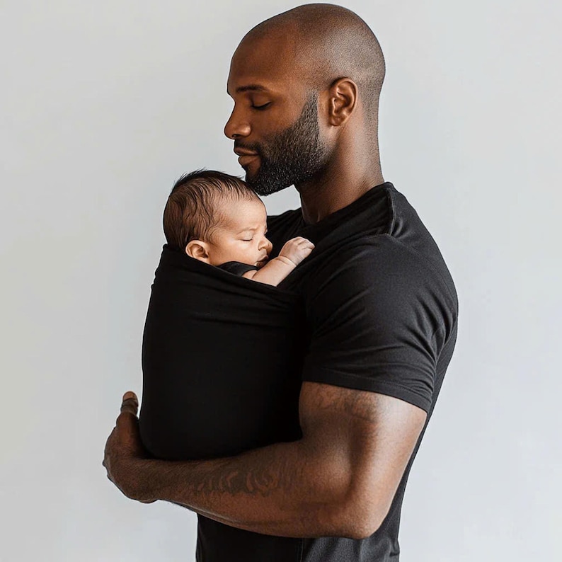 Peut inclure: Un porte-b&eacute;b&eacute; noir enveloppe un nourrisson endormi. Le porte-b&eacute;b&eacute; est tenu par une personne portant un t-shirt noir. L'arri&egrave;re-plan est un mur blanc uni.