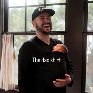 Peut inclure: T-shirt noir &agrave; manches longues et col en V avec un porte-b&eacute;b&eacute; int&eacute;gr&eacute;. Le texte "The dad shirt" est imprim&eacute; sur le t-shirt. Une casquette de baseball noire avec le mot "Dad" est port&eacute;e.