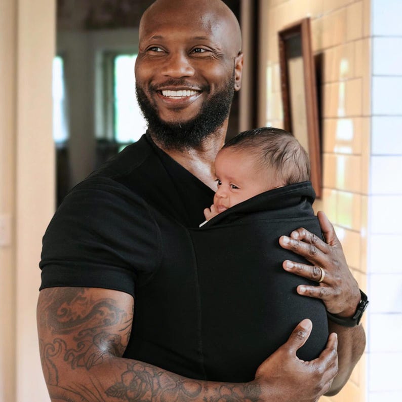 Peut inclure: Un porte-b&eacute;b&eacute; noir maintient un nourrisson. Le porte-b&eacute;b&eacute; est port&eacute; par une personne v&ecirc;tue d'un t-shirt noir. La personne a le cr&acirc;ne ras&eacute;, une barbe et des tatouages sur le bras. La personne sourit.