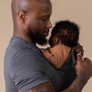 Peut inclure: Un porte-b&eacute;b&eacute; gris maintient un b&eacute;b&eacute; v&ecirc;tu d'un body orange. Le b&eacute;b&eacute; est tenu par une personne portant un t-shirt gris. La personne a une barbe et un tatouage sur le bras. L'arri&egrave;re-plan est de couleur beige uni.