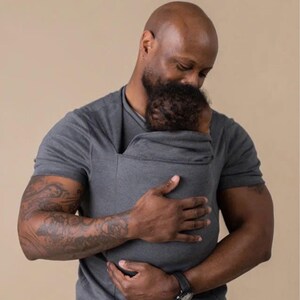 Peut inclure: Un porte-b&eacute;b&eacute; gris maintient un b&eacute;b&eacute; pr&egrave;s d'une personne. Le porte-b&eacute;b&eacute; est fait d'un mat&eacute;riau souple et flexible. La personne porte un t-shirt gris et a un tatouage sur le bras. Le fond est de couleur beige uni.