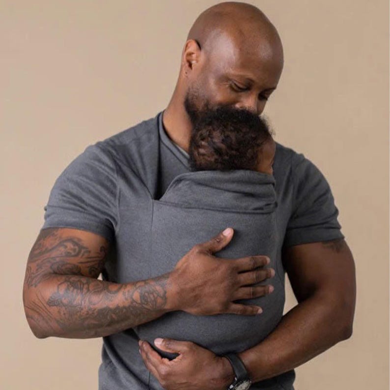 Peut inclure: Un porte-b&eacute;b&eacute; gris maintient un b&eacute;b&eacute; pr&egrave;s d'une personne. Le porte-b&eacute;b&eacute; est fait d'un mat&eacute;riau souple et flexible. La personne porte un t-shirt gris et a un tatouage sur le bras. Le fond est de couleur beige uni.