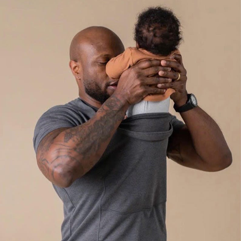 Peut inclure: Un porte-b&eacute;b&eacute; gris fonc&eacute; avec une poche int&eacute;gr&eacute;e, port&eacute; par une personne. Le b&eacute;b&eacute; porte un haut orange et un pantalon gris. La personne porte un t-shirt gris et une montre noire.