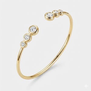 May include: A gold-colored open cuff bracelet with five round, clear gemstones. The gemstones are set in individual bezels and are evenly spaced along the top of the bracelet. The bracelet has a smooth, polished finish and a minimalist design.