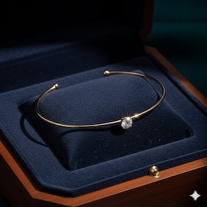 May include: A gold-colored bracelet with a single, clear, round gemstone set in the center. The open cuff design features small, rounded ends. The bracelet rests inside a dark blue velvet-lined jewelry box with a wooden exterior.