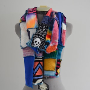 May include: A colorful patchwork scarf displayed on a mannequin. The scarf features a variety of knitted and sewn fabrics in vibrant colors, including blue, pink, yellow, and orange. Some sections have geometric patterns and a skull design.
