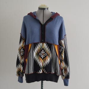 May include: A blue hooded sweater with a geometric pattern. The sweater features a color block design with a patterned front panel in black, white, yellow, and red. The sleeves and hood are solid blue with a burgundy trim.