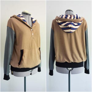 May include: A tan hoodie with gray sleeves and a striped hood lining. The hoodie has a front pocket and a black hem and cuffs. The front features three buttons. The back of the hoodie is also visible.