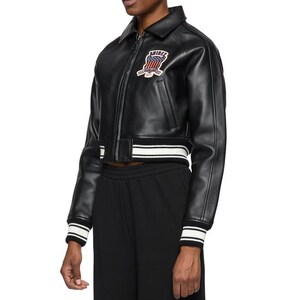 K&ouml;nnte beinhalten: Schwarze Leder-Bomberjacke mit Rei&szlig;verschluss, Kragen und Aufn&auml;her auf der Brust. Die Jacke hat wei&szlig;-schwarze Streifen an den Manschetten und am Saum. Der Aufn&auml;her zeigt das Wort "AMIRI".