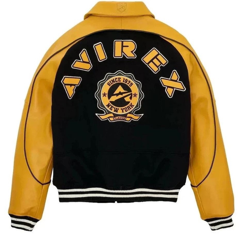Puede incluir: Chaqueta estilo universitario en negro y amarillo mostaza. La parte trasera de la chaqueta presenta la palabra "AVIREX" en letras amarillas, y un emblema circular con el texto "SINCE 1975 NEW YORK CLASSIC". Los pu&ntilde;os tienen rayas blancas y negras.