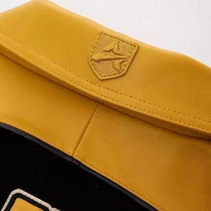 Puede incluir: Detalle de una chaqueta de cuero amarillo mostaza con una secci&oacute;n inferior negra. La chaqueta presenta un peque&ntilde;o emblema bordado en la parte superior derecha. El emblema tiene forma de escudo con un dise&ntilde;o estilizado.