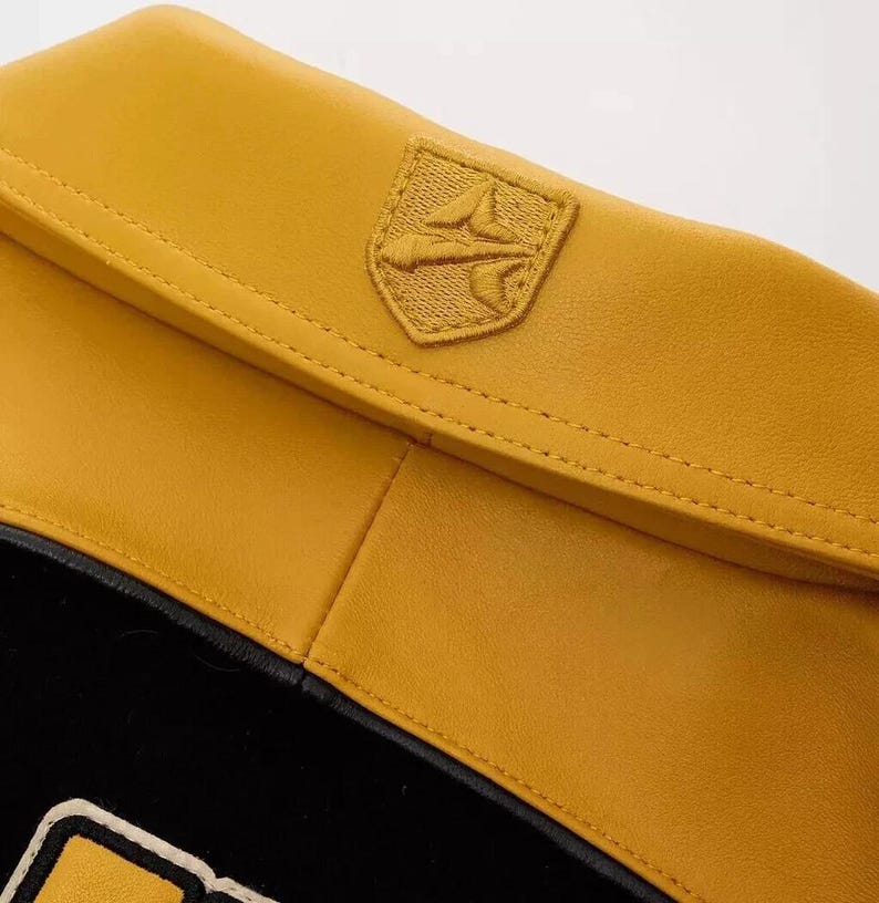 Puede incluir: Detalle de una chaqueta de cuero amarillo mostaza con una secci&oacute;n inferior negra. La chaqueta presenta un peque&ntilde;o emblema bordado en la parte superior derecha. El emblema tiene forma de escudo con un dise&ntilde;o estilizado.