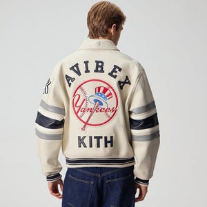 Könnte beinhalten: Cremefarbene Lederjacke mit den Logos "AVIREX" und "Yankees" auf dem Rücken. Die Jacke hat marineblaue und graue Streifen auf den Ärmeln und den Text "KITH" am unteren Rand. Die Jacke hat eine Knopfleiste und einen Kragen.