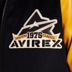 Puede incluir: Primer plano de una chaqueta universitaria negra y amarilla. La chaqueta presenta un parche con el texto "NEW YORK 1975 CLASSIC AVIREX" en blanco, negro y amarillo. La chaqueta tiene un cuerpo negro y mangas amarillas.