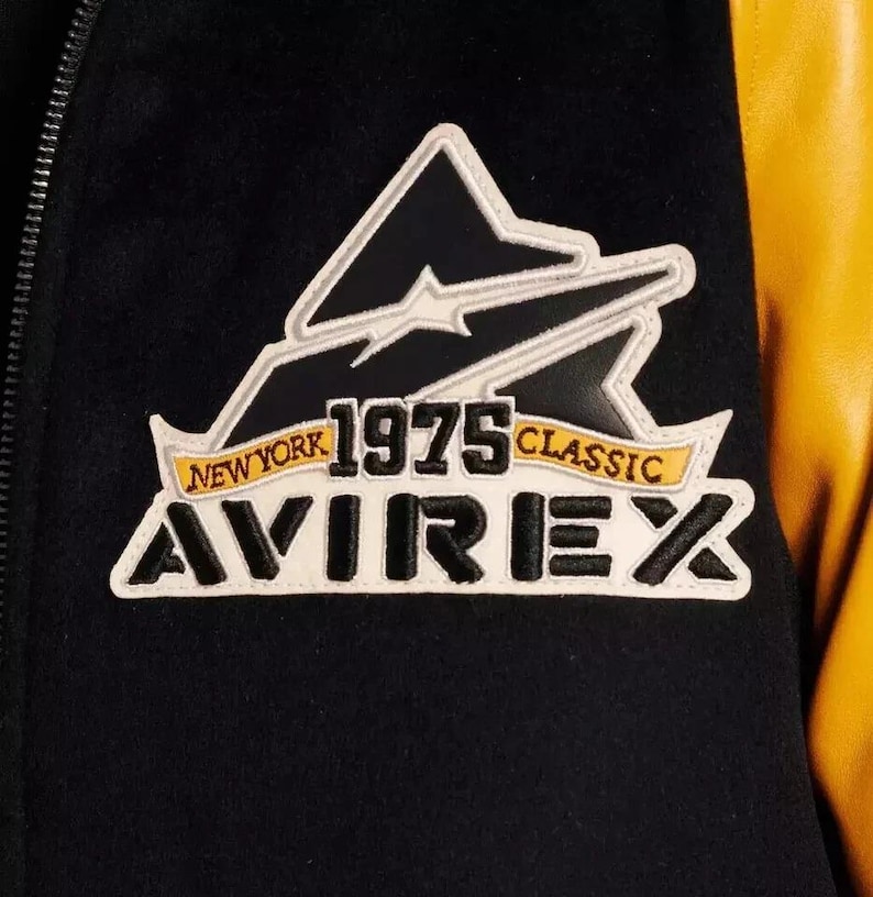 Puede incluir: Primer plano de una chaqueta universitaria negra y amarilla. La chaqueta presenta un parche con el texto "NEW YORK 1975 CLASSIC AVIREX" en blanco, negro y amarillo. La chaqueta tiene un cuerpo negro y mangas amarillas.