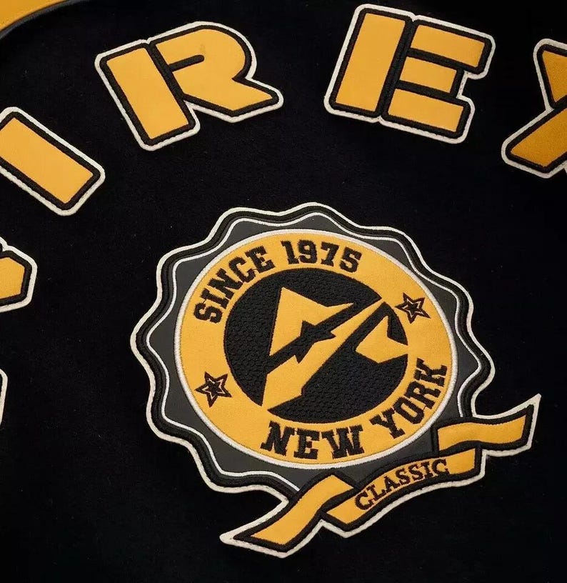 Puede incluir: Tela negra con parches bordados amarillos y blancos. Los parches incluyen la palabra "REX" en letras amarillas y un emblema circular con el texto "SINCE 1975 NEW YORK CLASSIC".