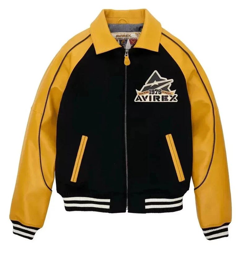 Puede incluir: Chaqueta universitaria negra y amarilla con el texto "AVIREX NEW YORK 1975 CLASSIC" en el pecho. La chaqueta tiene cuello, mangas y ribetes de bolsillo amarillos, con cuerpo negro y pu&ntilde;os y dobladillo a rayas blancas.