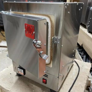 May include: A stainless steel kiln with a red logo on the door. The kiln has a silver handle and a red power button. The interior is lined with a beige material. A black power cord is visible.