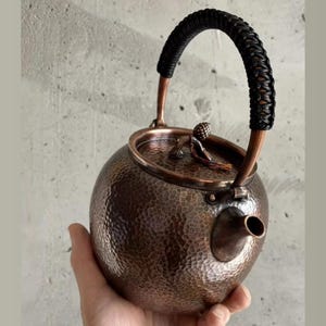 Könnte beinhalten: Eine Kupfer-Teekanne mit gehämmertem Muster, mit einem schwarzen geflochtenen Griff und einem kleinen Ausguss. Der Deckel hat einen dekorativen Knopf. Die Teekanne wird in einer Hand gehalten.