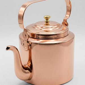Könnte beinhalten: Ein kupferfarbener Teekessel mit gebogenem Ausguss und großem, gewölbtem Griff. Der Deckel ist mit einem kleinen, goldfarbenen Knopf versehen. Der Kessel hat eine polierte, reflektierende Oberfläche und ist für den Gebrauch auf dem Herd konzipiert.