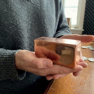 May include: A rectangular copper bar with a polished finish is held in open hands. The bar reflects the surrounding environment. The person holding the bar is wearing a gray sweater. The bar's dimensions are not visible.