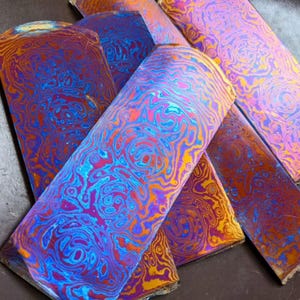 Può includere: Primo piano di diversi pezzi rettangolari di metallo con un motivo vorticoso vibrante. I colori includono sfumature di blu, viola, arancione e oro, creando un aspetto artistico unico. I pezzi sembrano essere tagliati da un foglio più grande.