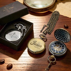 Puede incluir: Tres relojes de bolsillo se muestran sobre una superficie de madera. Uno está en una caja negra, grabado con "MICHAEL". Otro está abierto, mostrando la hora. El tercero está cerrado, grabado con "Jason Thomas". Una cadena está unida a un reloj.