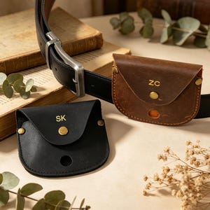 Puede incluir: Bolsas de cinturón de cuero negro y marrón con iniciales. La bolsa negra tiene las iniciales "SK" y la bolsa marrón tiene las iniciales "ZC". Ambas bolsas están unidas a un cinturón de cuero negro con una hebilla plateada. Las bolsas están aseguradas con remaches de color latón.