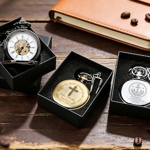 Op de afbeelding: Drie zakhorloges in zwarte geschenkdozen. Eén horloge heeft een open wijzerplaat met een witte achtergrond en een goudkleurig skeletuurwerk. De andere twee zijn gesloten, één goudkleurig met een kruis en tekst, één zilverkleurig met een anker en tekst.