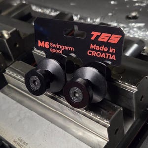 May include: Two black M6 swingarm spools with a silver center, displayed on a metal surface. The product packaging is black and red, with the text "TSS Made in CROATIA".