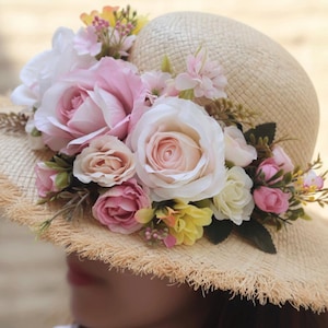 Könnte beinhalten: Ein Sonnenhut aus Stroh in Beige, verziert mit rosa, weißen und gelben Rosen. Der Hut hat eine ausgefranste Krempe und ist mit verschiedenen floralen Akzenten verziert. Der Hut hat eine hellbeige Farbe.