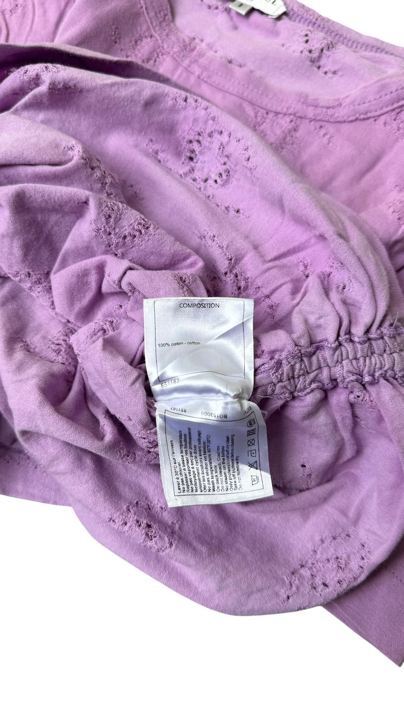 May include: A light purple, short-sleeved shirt with a textured design and a gathered waist. The shirt has a tag that reads "COMPOSITION" and "100% coton / cotton".