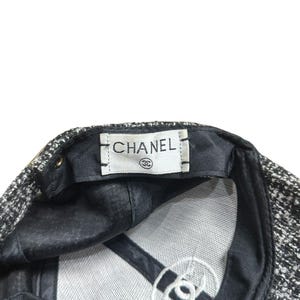 Op de afbeelding: Een zwart-witte tweed baseballcap met de merknaam "CHANEL" gedrukt op een wit label. De binnenkant heeft een zwarte voering en een wit geborduurd logo.