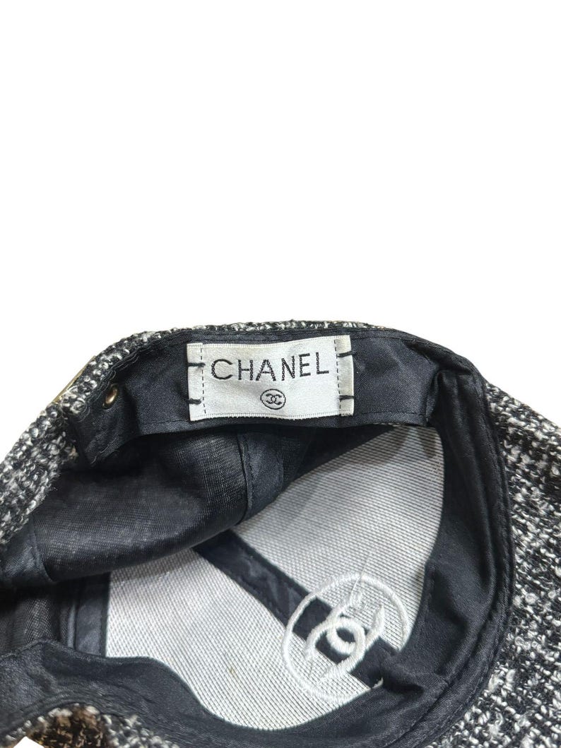 Op de afbeelding: Een zwart-witte tweed baseballcap met de merknaam "CHANEL" gedrukt op een wit label. De binnenkant heeft een zwarte voering en een wit geborduurd logo.