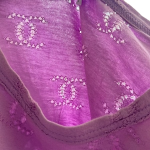 May include: A close-up of a lavender-colored top with a repeating pattern of interlocking Cs. The top has a scoop neck and a tag that reads "CHANEL". The fabric appears to be lightweight and semi-sheer.