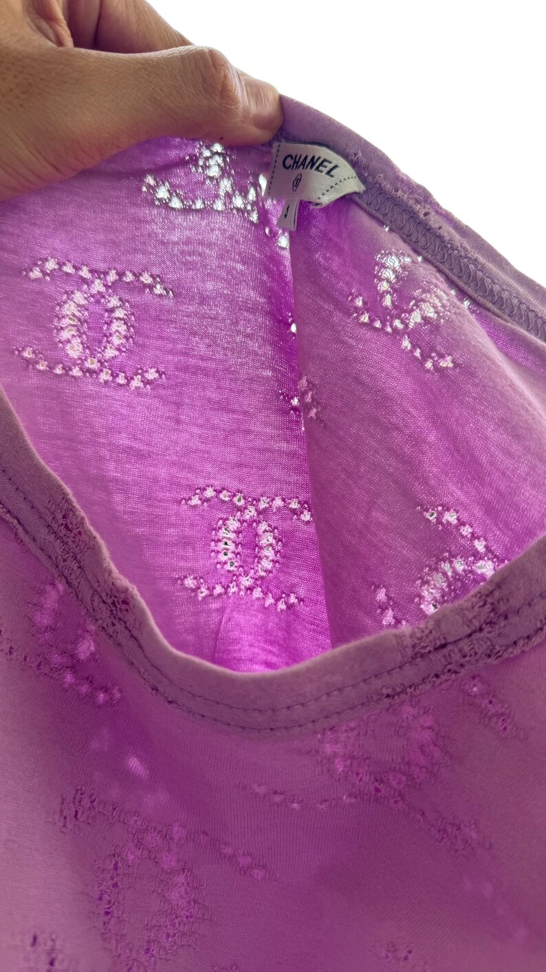 May include: A close-up of a lavender-colored top with a repeating pattern of interlocking Cs. The top has a scoop neck and a tag that reads "CHANEL". The fabric appears to be lightweight and semi-sheer.