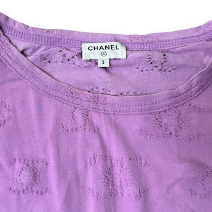 May include: A lilac-colored, short-sleeved shirt with a crew neck. The shirt features a repeating pattern of the Chanel logo and the Chanel label is visible at the neckline. The shirt is size small.