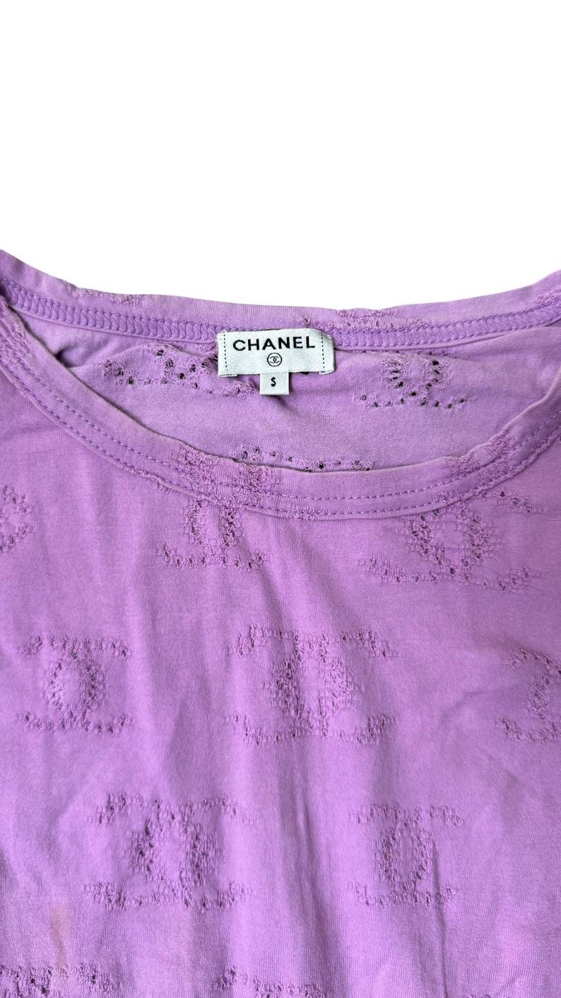 May include: A lilac-colored, short-sleeved shirt with a crew neck. The shirt features a repeating pattern of the Chanel logo and the Chanel label is visible at the neckline. The shirt is size small.