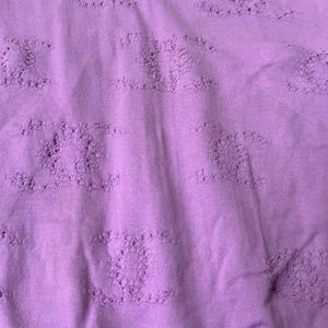 May include: A lilac-colored t-shirt with a crew neck and short sleeves. The shirt features a textured pattern throughout and a small label with the brand name "CHANEL" at the neckline.