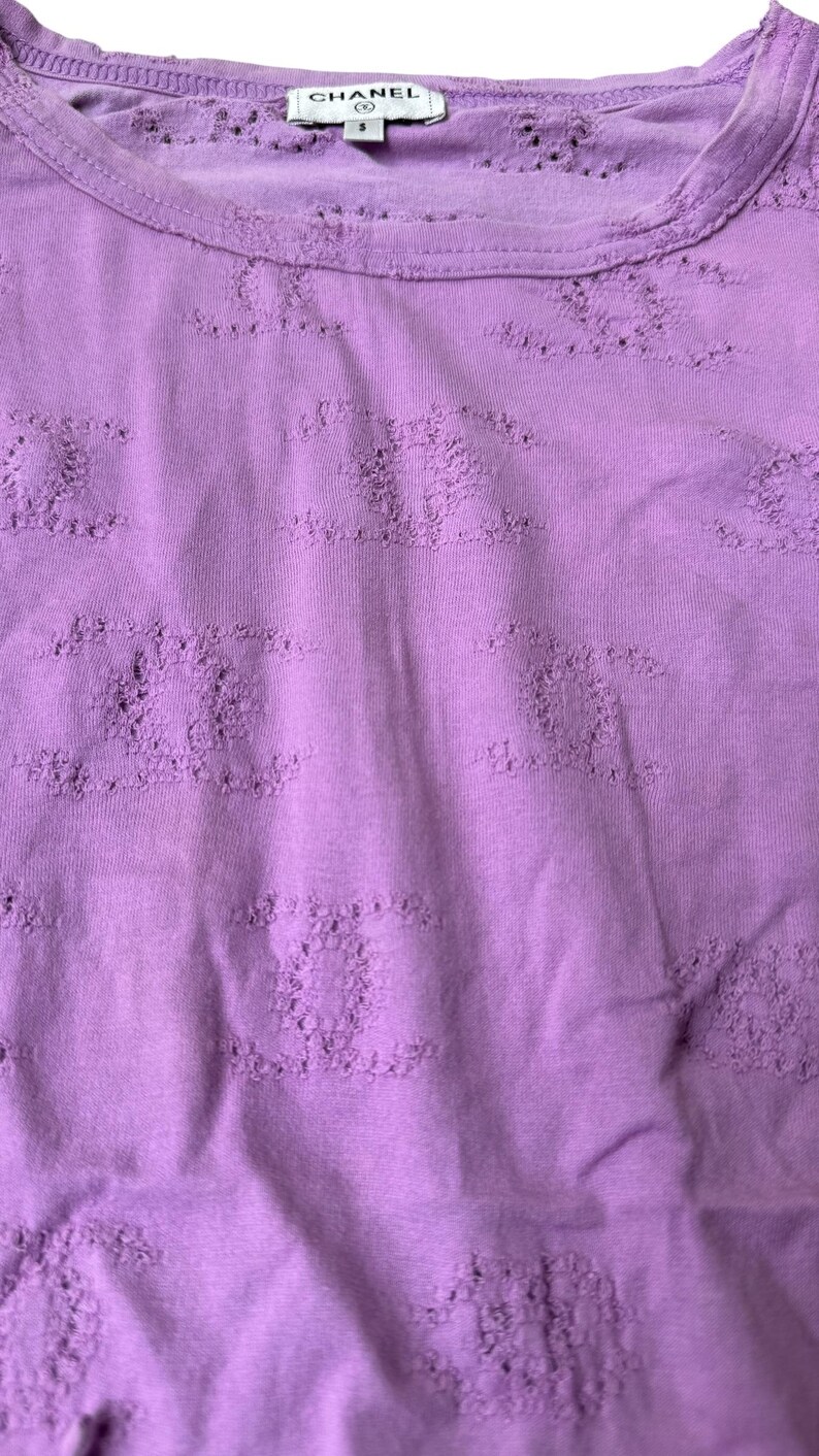May include: A lilac-colored t-shirt with a crew neck and short sleeves. The shirt features a textured pattern throughout and a small label with the brand name "CHANEL" at the neckline.