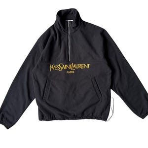 May include: Black fleece pullover with a stand-up collar and a quarter-zip. The front displays gold text reading "Yves Saint Laurent Paris". It has long sleeves, a front pocket, and a drawstring hem.